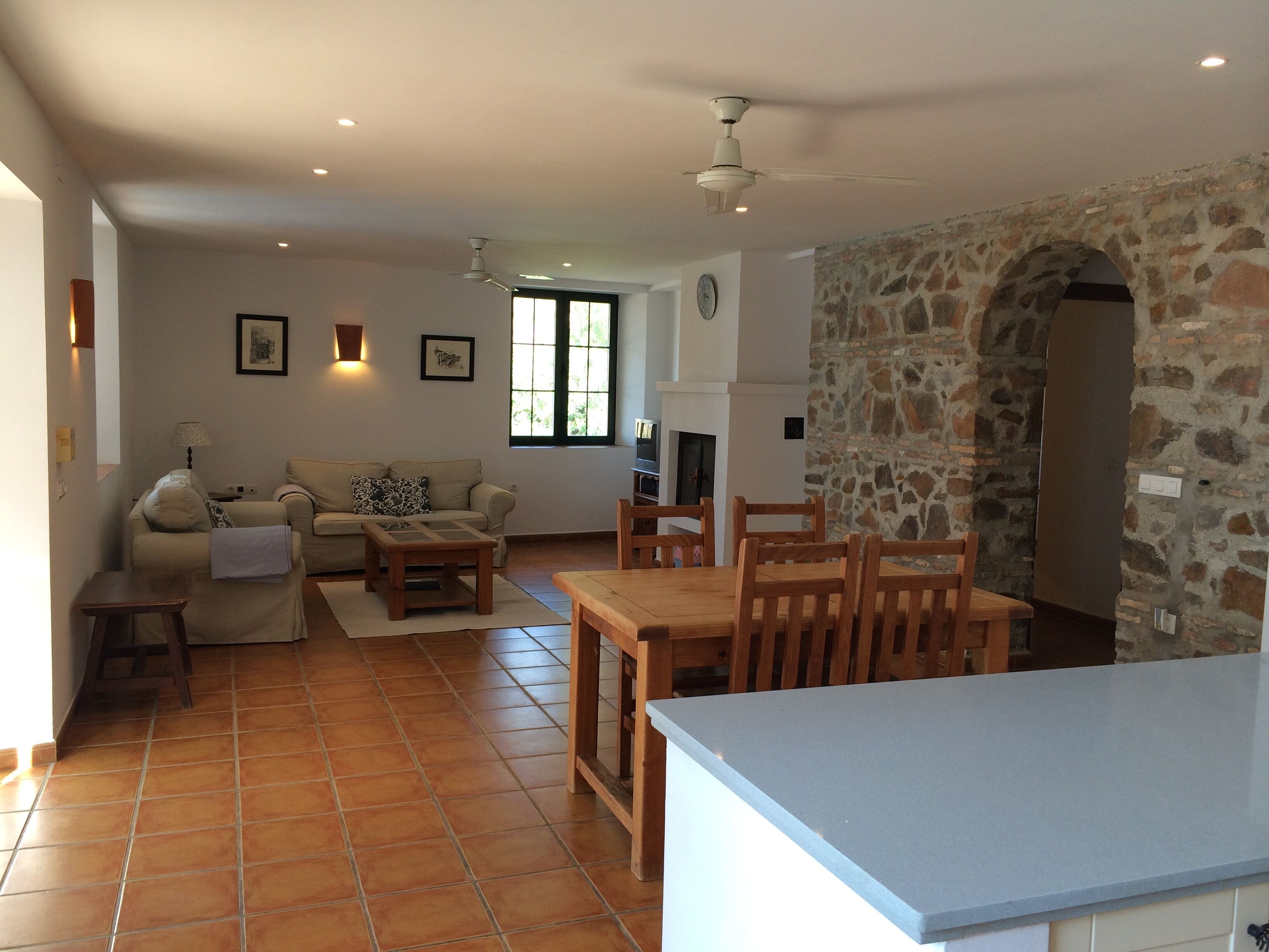 Un'ampia zona giorno con divano, tavolo da pranzo con sedie, angolo cottura con parete in pietra e ventilatore a soffitto.