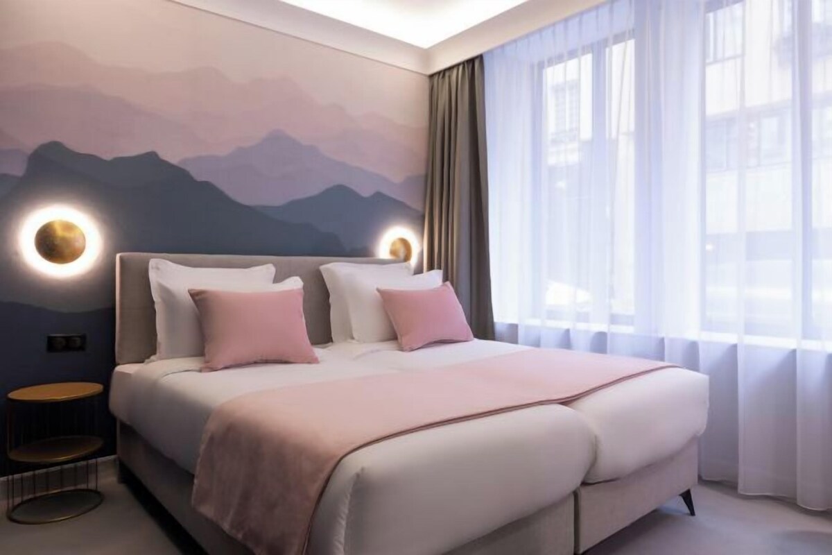 A modern bedroom with a large bed, a wall-mounted light, and a view of the outdoors.