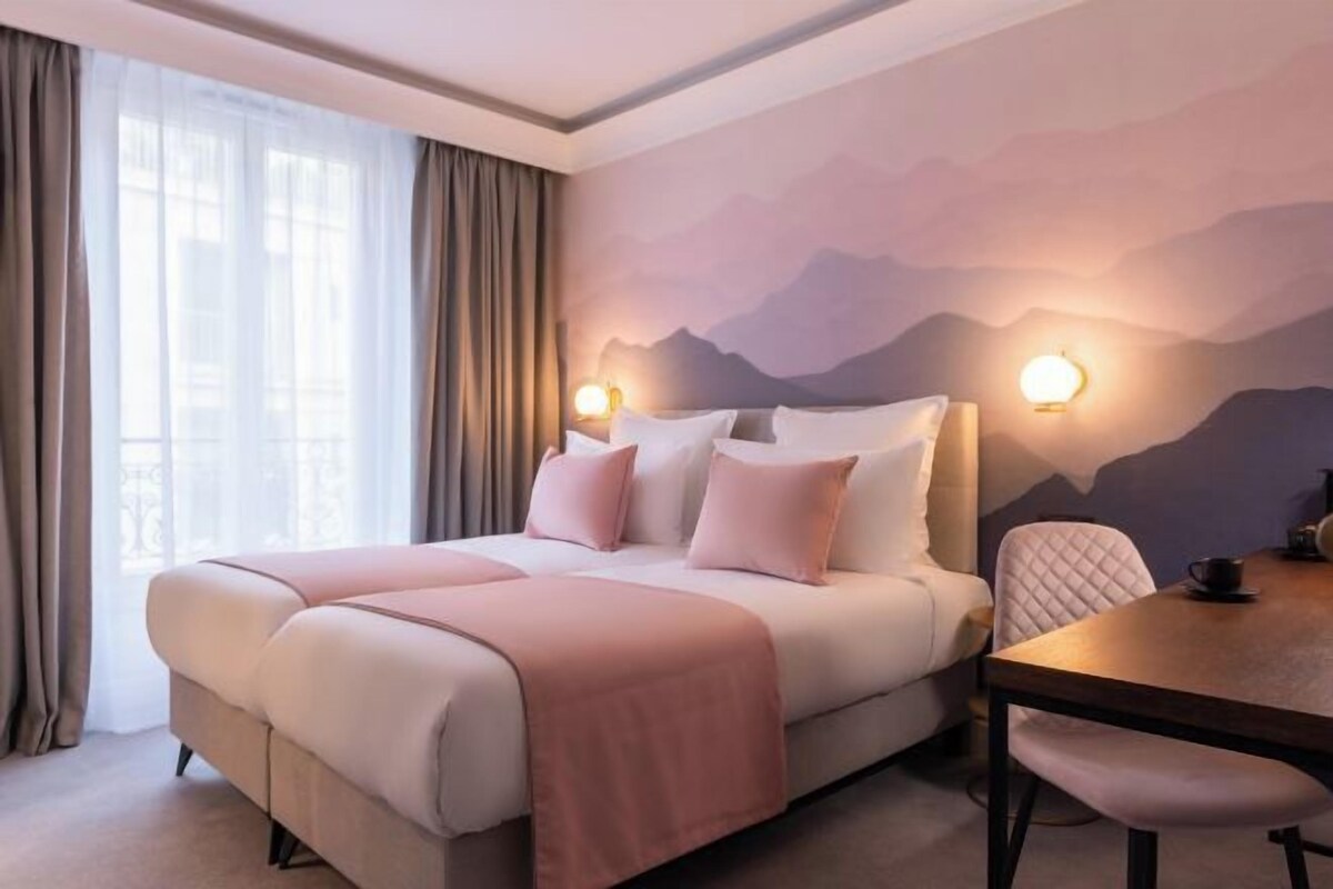 Habitación de hotel con cama, una mesa y sillas. Se ve una ventana con cortinas, un muro con un mural de montaña y dos lámparas de pared.