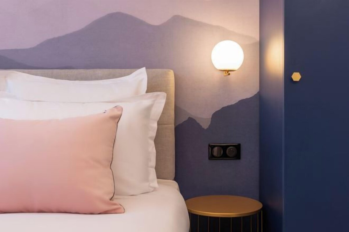 A bedroom with a bed, pillows, a round side table, and a wall-mounted light fixture.