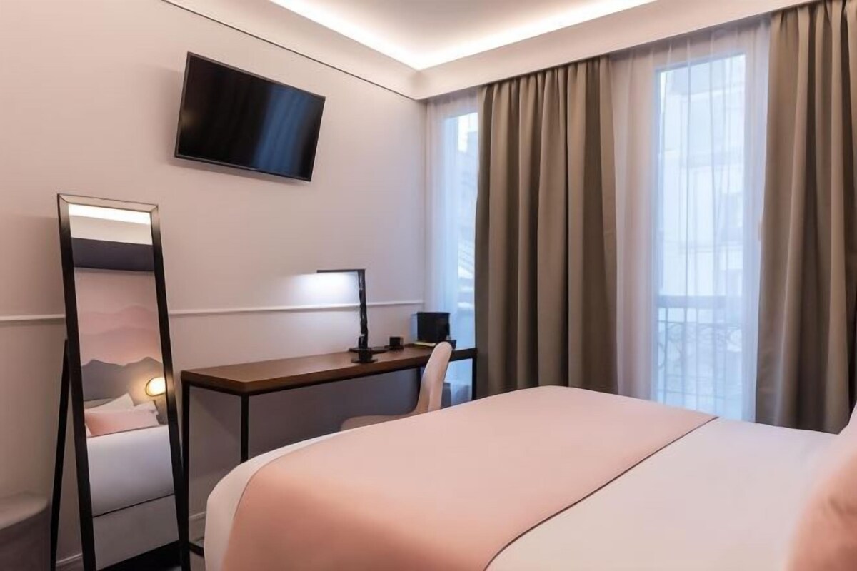 Habitación de hotel con cama, escritorio, silla, televisor y ventana grande con cortinas.