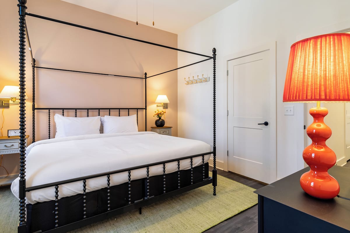 A four-poster bed with white linens, a nightstand with a lamp, and a red lamp on a table.