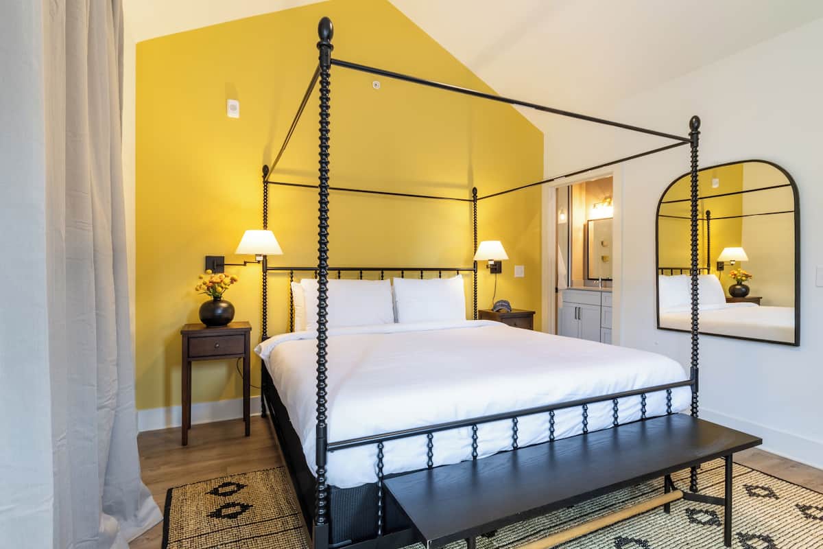 A four-poster bed with white linens, a bedside table with a lamp, and a mirror on the wall.