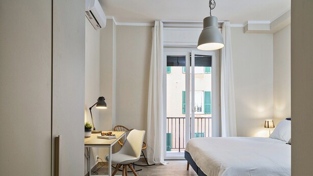 Um quarto com cama, escrivaninha com cadeira, luminária e vista para o exterior.