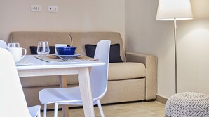 Uma sala de estar aconchegante com um sofá bege, uma mesa branca com uma tigela azul, duas cadeiras brancas, um abajur e um banquinho de tricô.