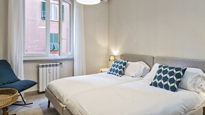 Quarto com cama, duas almofadas, uma cadeira, abajur e janela com persianas.