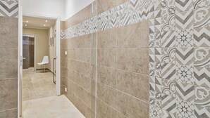 Um corredor com ladrilhos estampados, uma porta que dá para um banheiro e uma cadeira branca.
