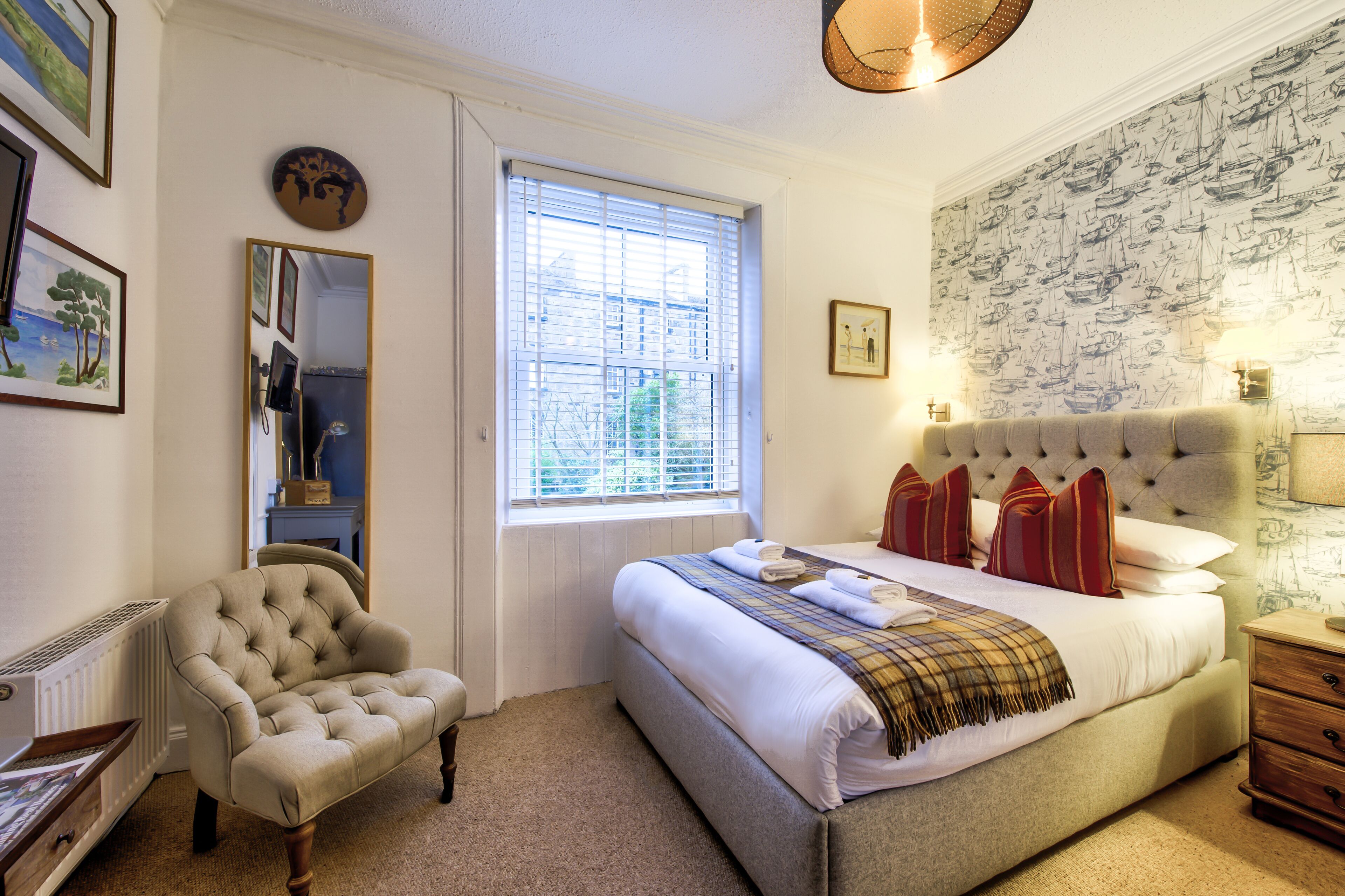 A bedroom with a bed, a chair, a window, a mirror, and a wall with nautical wallpaper.