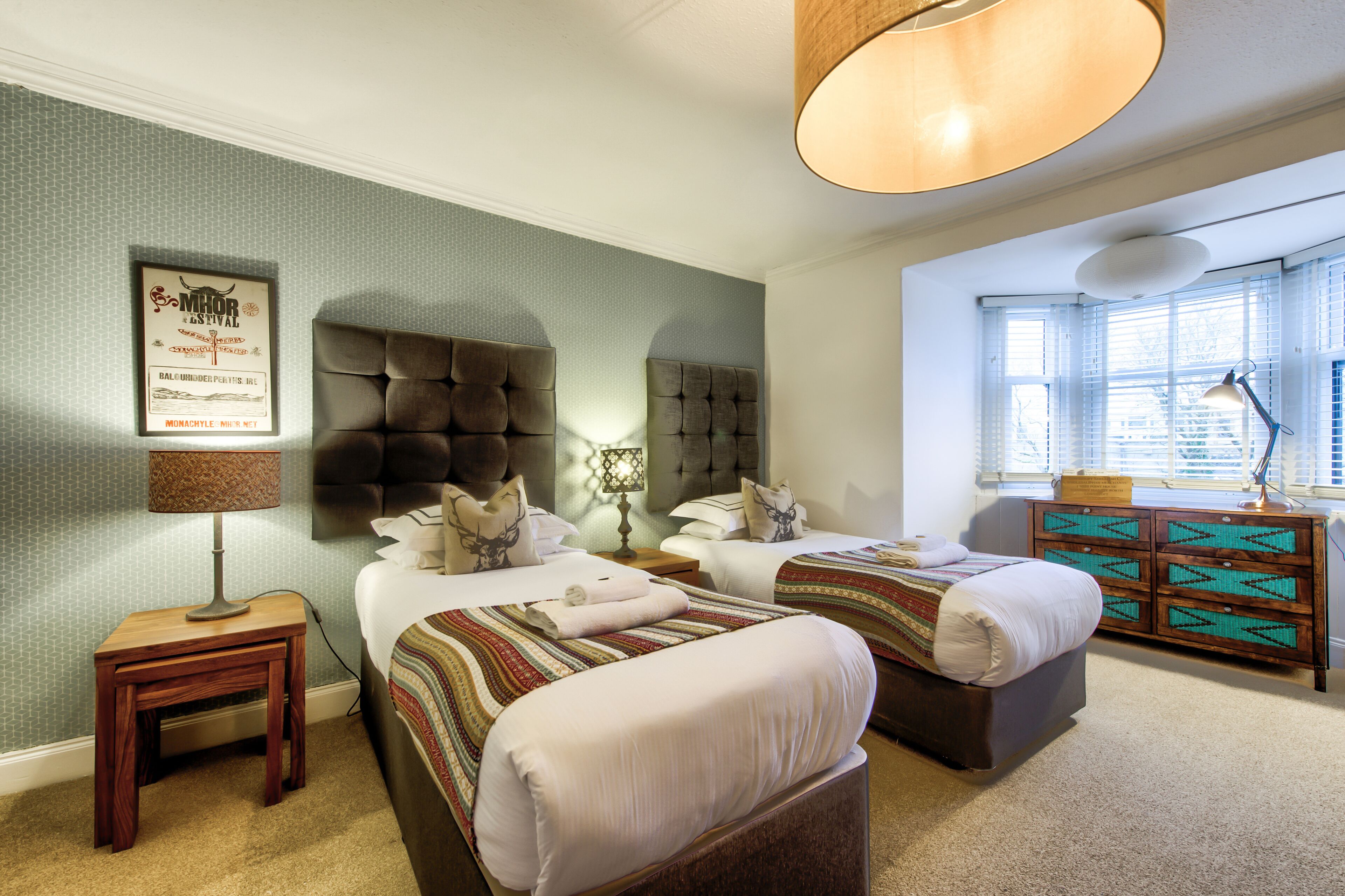 A hotel room with two beds, a wooden nightstand with a lamp, a decorative wall piece, and a window with blinds.