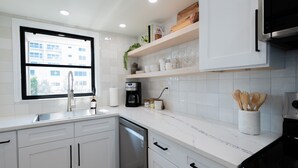A modern kitchen with white cabinetry, a built-in dishwasher, a coffee maker, and a window providing natural light. - Newly Reno Luxury Condo 30 Steps to Paradise (Clearwater Beach)
