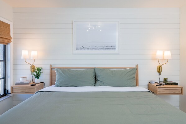 Ein ordentlich eingerichtetes Schlafzimmer mit einem großen Bett, zwei Nachttischen, einer Wandlampe und einem gerahmten Bild über dem Bett.