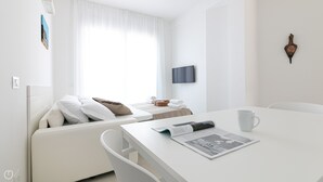 A modern, minimalist hotel room with a white color scheme, featuring a bed with white and beige pillows, a small table with a magazine and a white mug, and a television mounted on the wall. - Rivazzurra Homes - 13 (Termoli)