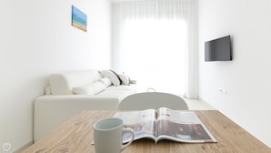A modern, minimalistic living room with a white sofa, a coffee table with a magazine, and a TV mounted on the wall. - Rivazzurra Homes - 20 (Termoli)