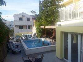 Zona de piscina con sillas de descanso y sombrillas, rodeada de follaje y edificios.