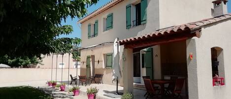 Un bâtiment à deux étages, doté d’un toit en tuiles, de volets verts et d’un patio couvert aménagé avec du mobilier d’extérieur.