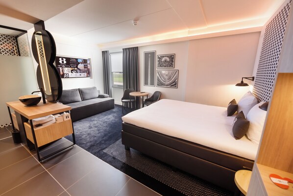 A modern hotel room with a large bed, a desk with a bowl, a sofa, a small table, and a wall-mounted TV. - Gr8 Hotel Maastricht-Aachen Airport (Maastricht-Airport)