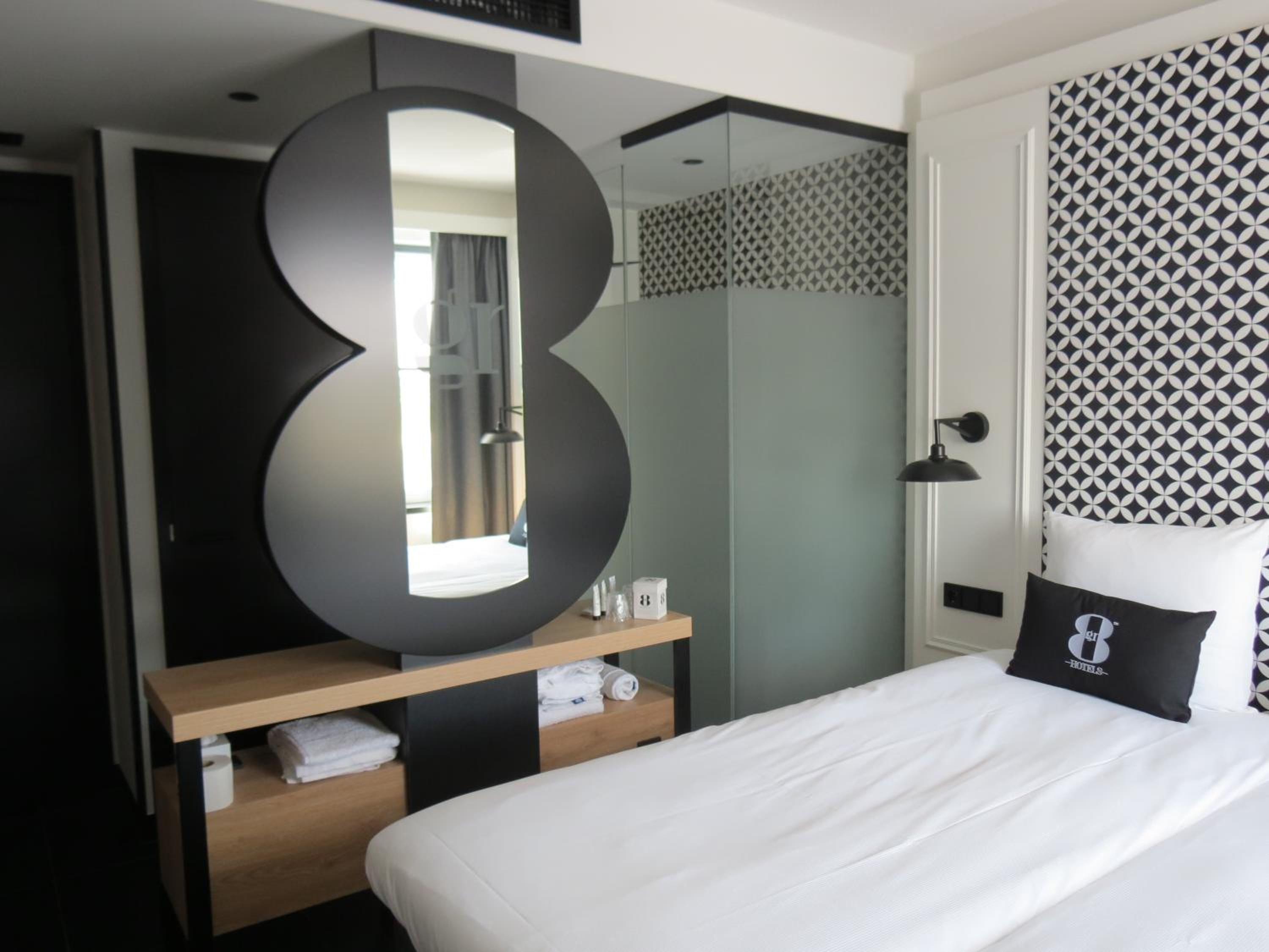 Une chambre d’hôtel moderne avec un grand miroir, un lit avec une literie blanche, une table de chevet en bois et un mur à motifs.
