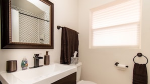 A bathroom with a white sink, dark wooden cabinet, a mirror, a towel rack, a toilet, and a window with blinds.
