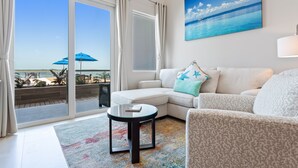 A modern living room with a white sofa, a patterned rug, and a glass coffee table. There is a large window with curtains, a painting of a beach, and a view of the ocean. - Sunset Caribe Belize (San Pedro)