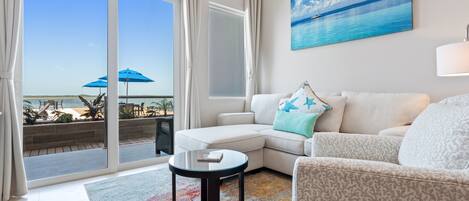 Una sala de estar moderna con un sofá blanco, una alfombra estampada y una mesa de centro de vidrio. Se ve una ventana grande con cortinas, un cuadro de una playa y vistas al océano.