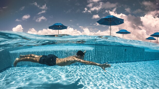 An underwater view of a swimming pool with a person swimming, clear blue water, and several blue umbrellas in the background. - Sunset Caribe Belize (San Pedro)