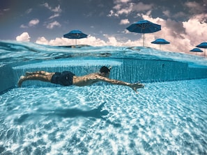 Vista subacuática de una piscina con una persona nadando, agua azul cristalina y varias sombrillas azules al fondo.