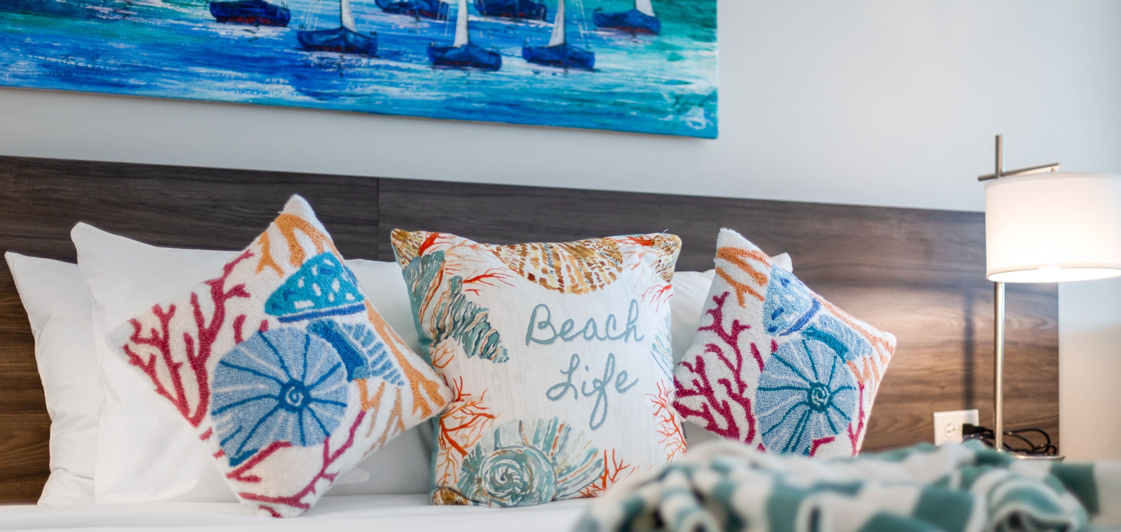 A bed with colorful pillows and a painting of boats on the wall.
