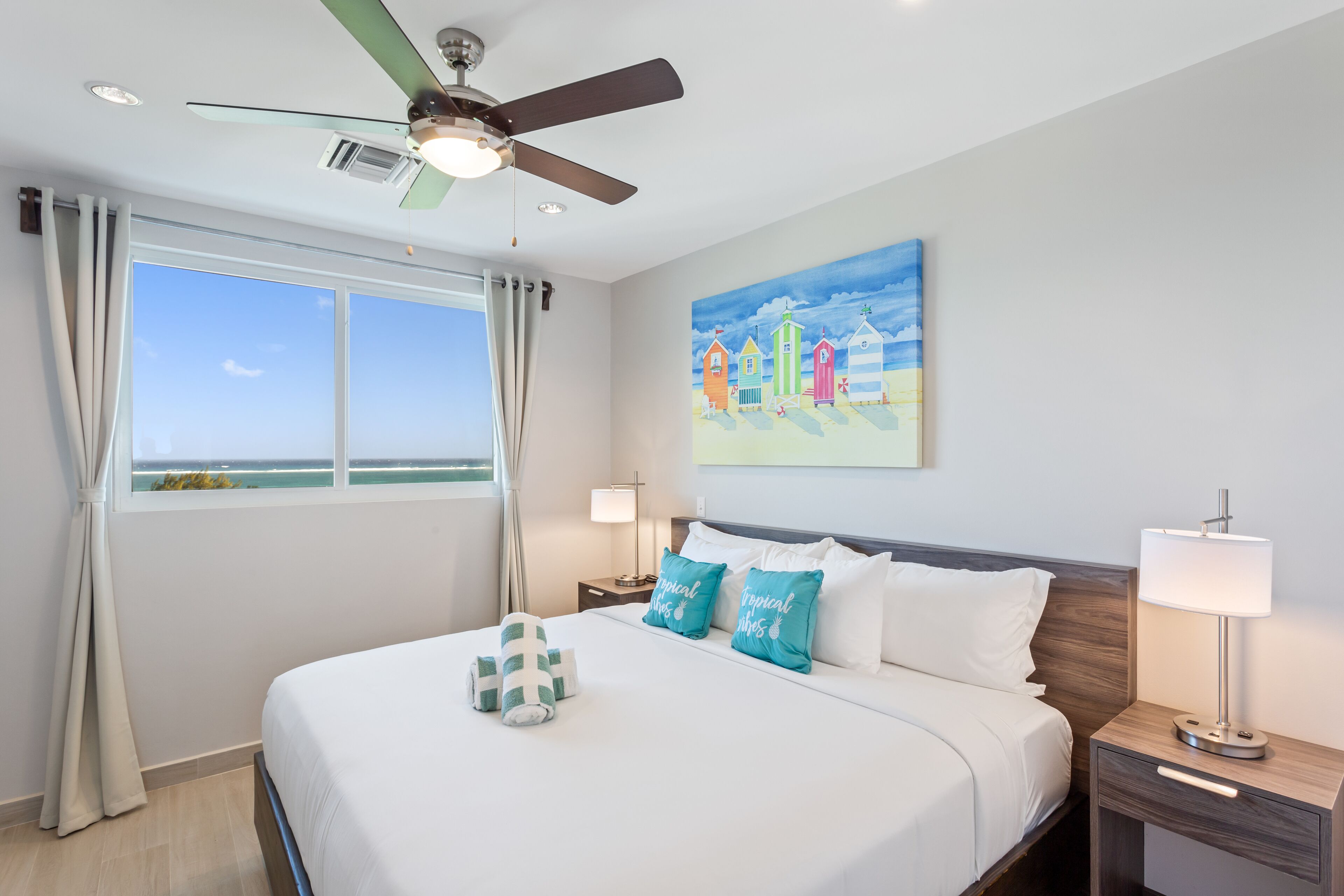 Una habitación de hotel con una cama, un ventilador de techo, una mesita de noche con una lámpara, un cuadro con casas de playa y una ventana con cortinas.