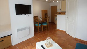 A compact living space with a flat-screen TV mounted on the wall, a dining area with wooden chairs and a table, and a kitchenette with a white cabinet. - Apartment sables d'or les pins (Fréhel)