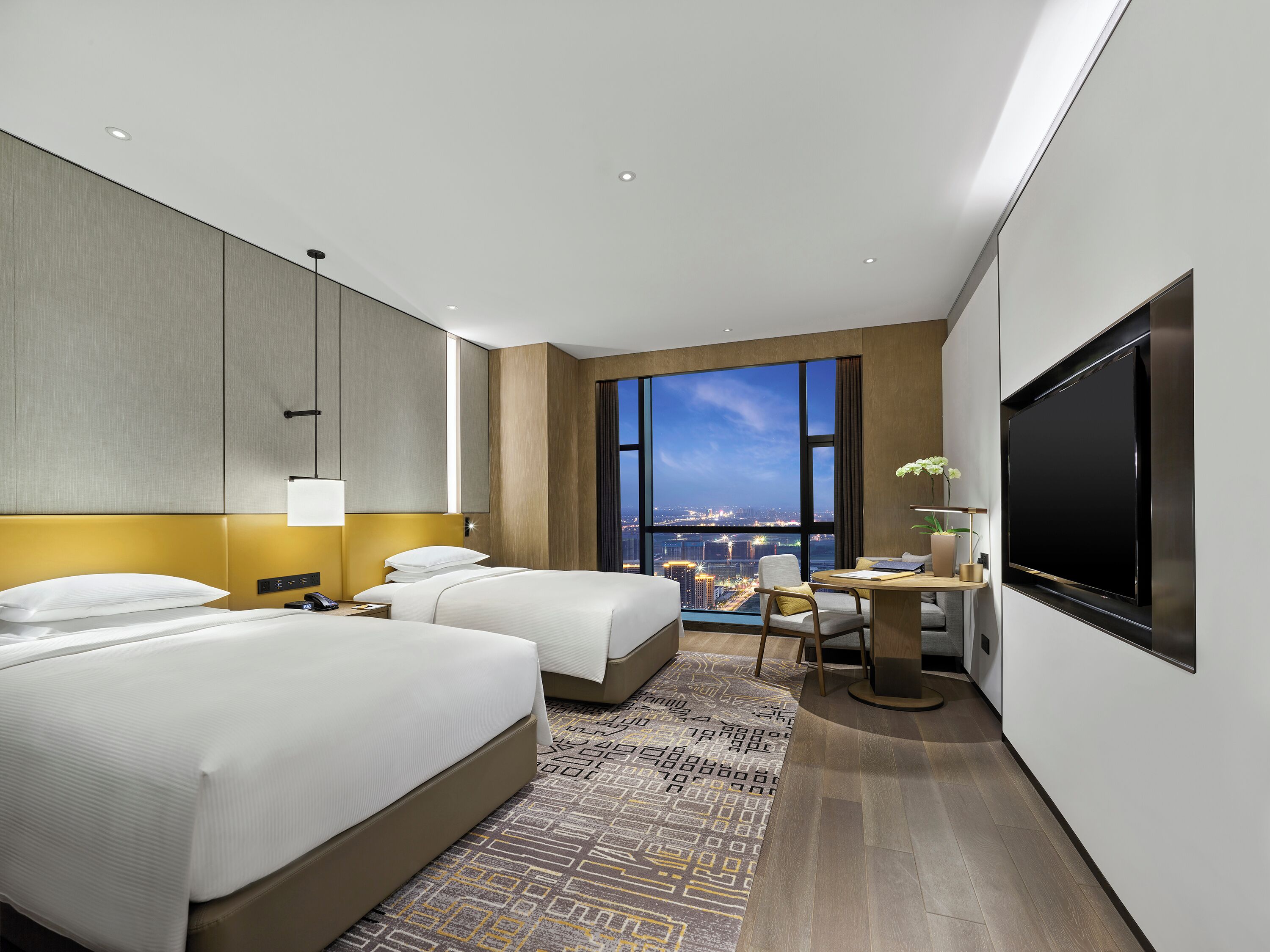 Habitación de hotel moderna con dos camas, un escritorio, un televisor y un amplio ventanal con vistas a la ciudad.