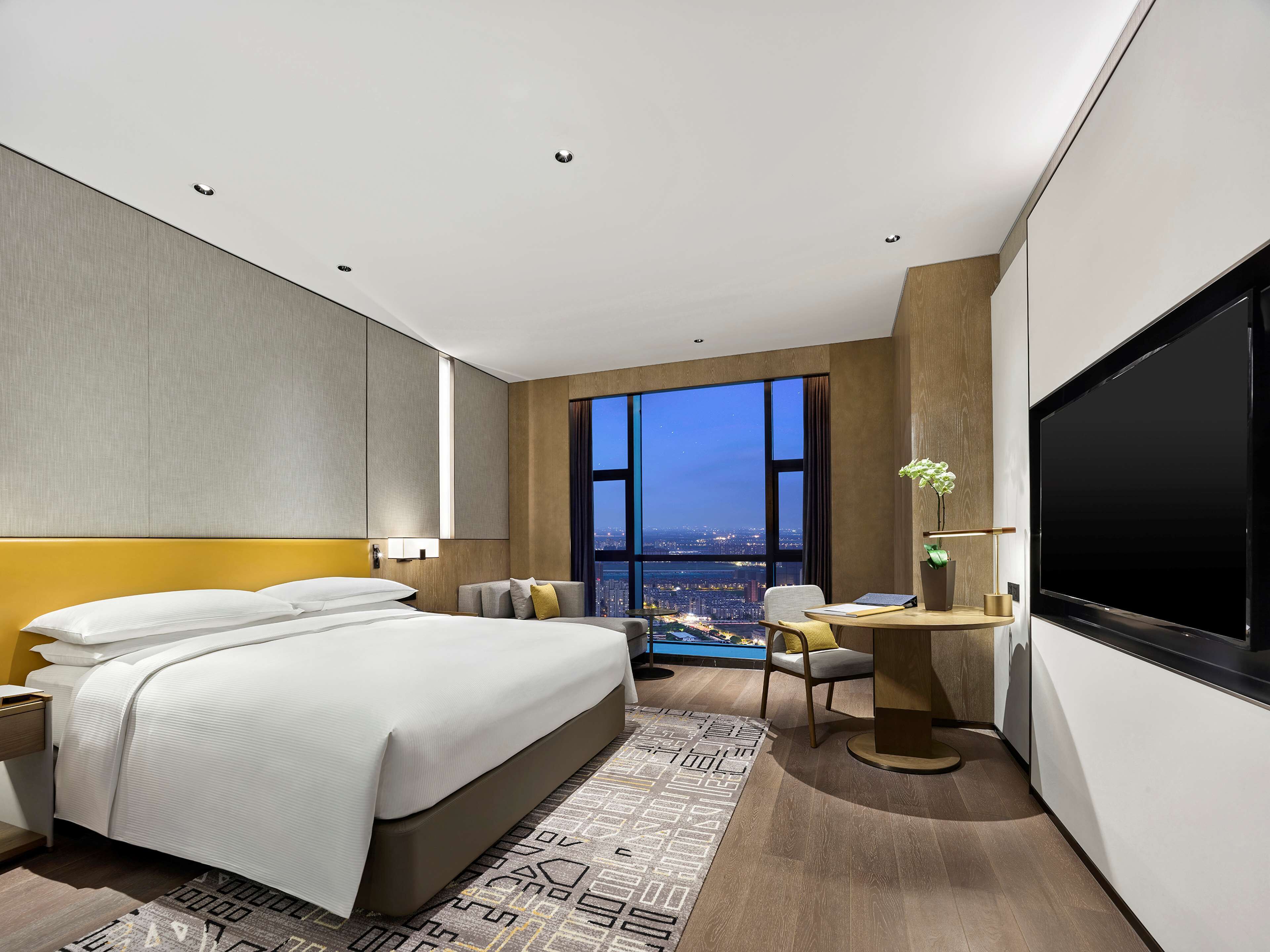 Una habitación de hotel moderna con una cama grande, un escritorio con una silla, un televisor de pantalla plana y vistas a la ciudad de noche.