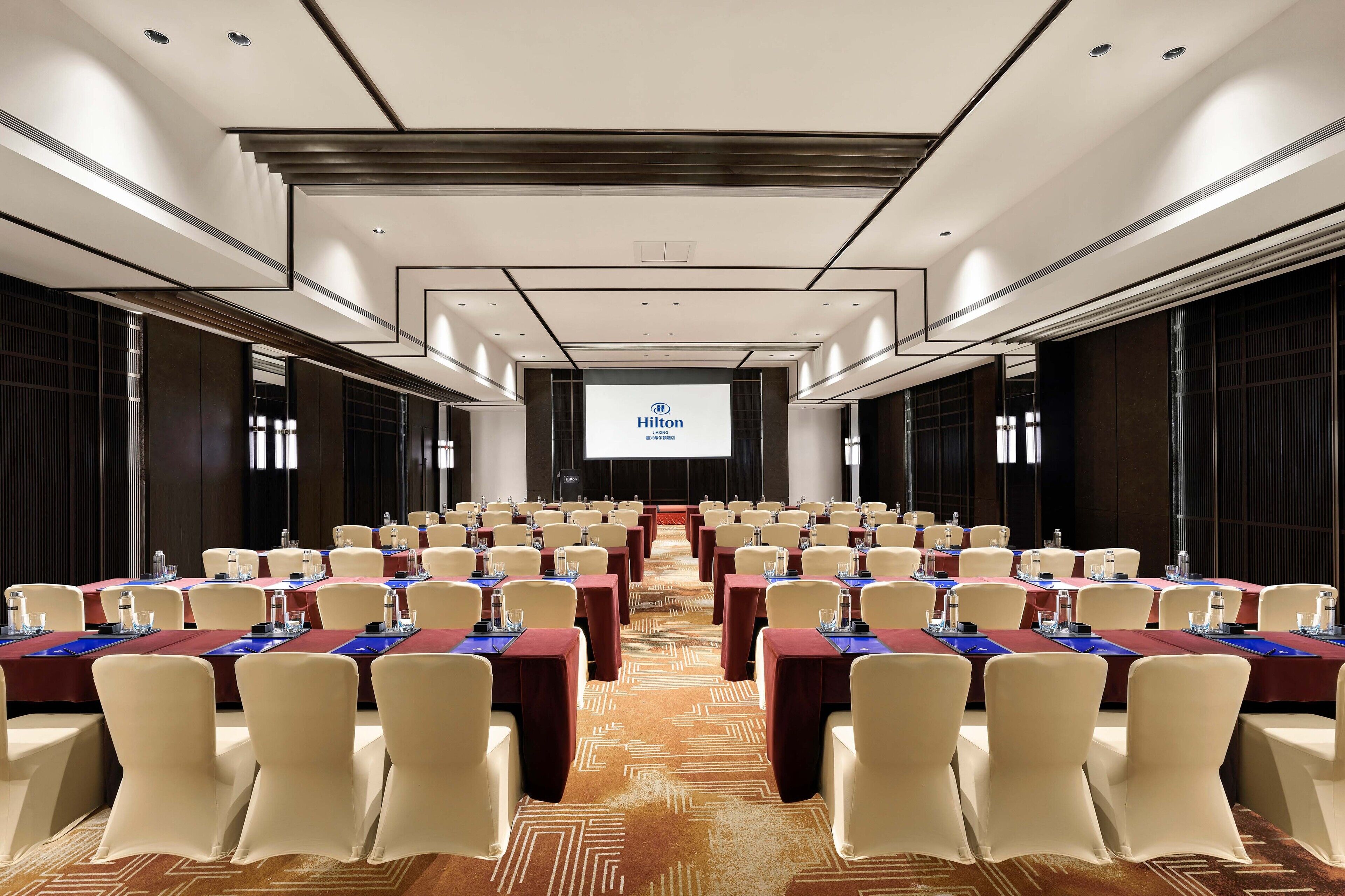 A conference room with rows of tables and chairs, a large screen at the front, and a carpeted floor.