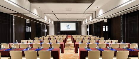 A conference room with rows of tables and chairs, a large screen at the front, and a carpeted floor.