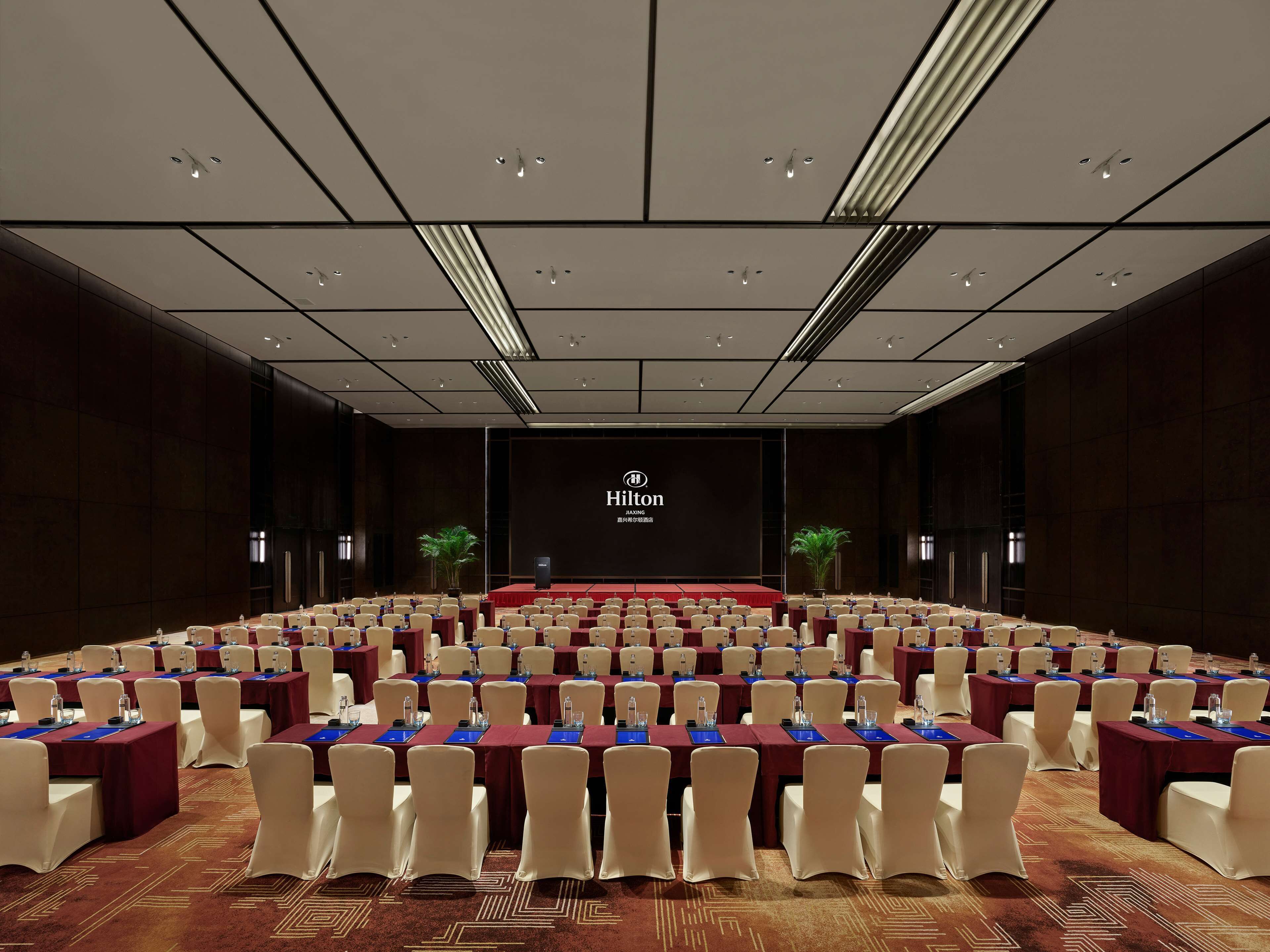Une grande salle de conférence avec des rangées de tables et de chaises, une scène ornée du logo « Hilton » et deux plantes en pot.