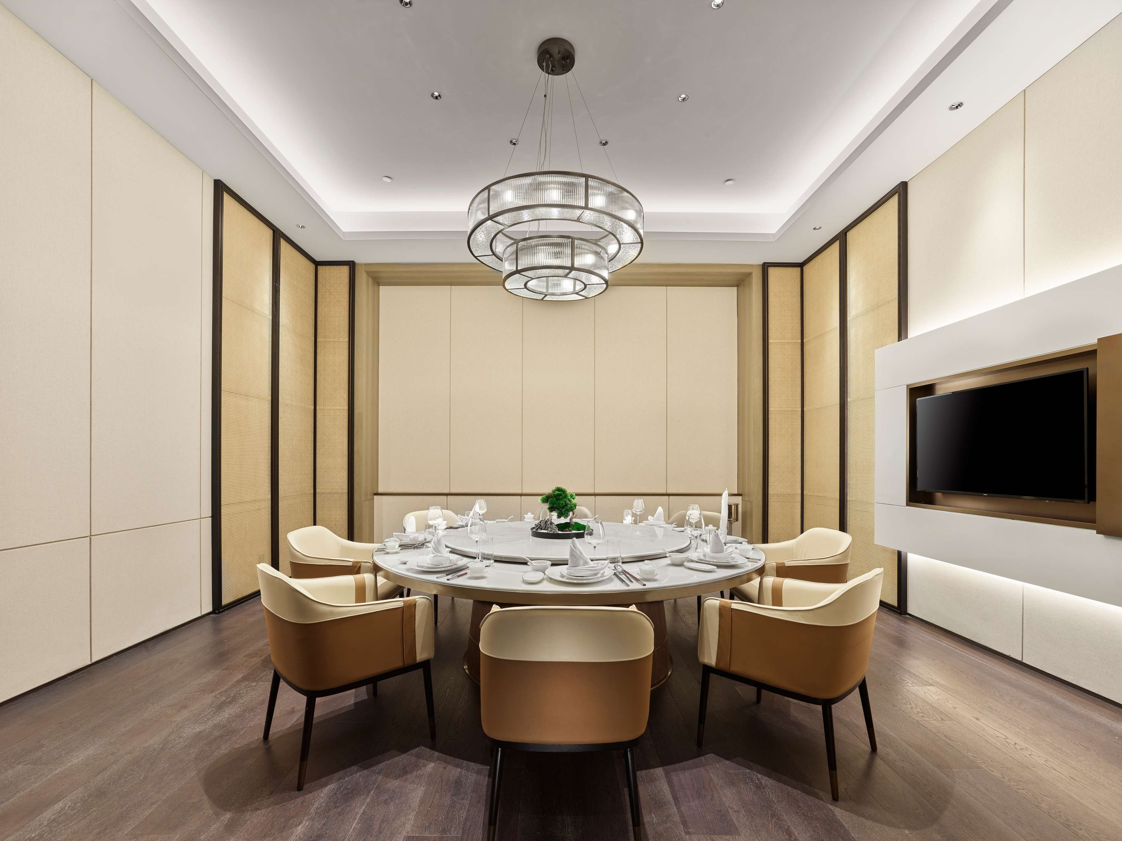 A modern conference room with a round table, six chairs, a chandelier, and a flat-screen TV mounted on the wall.