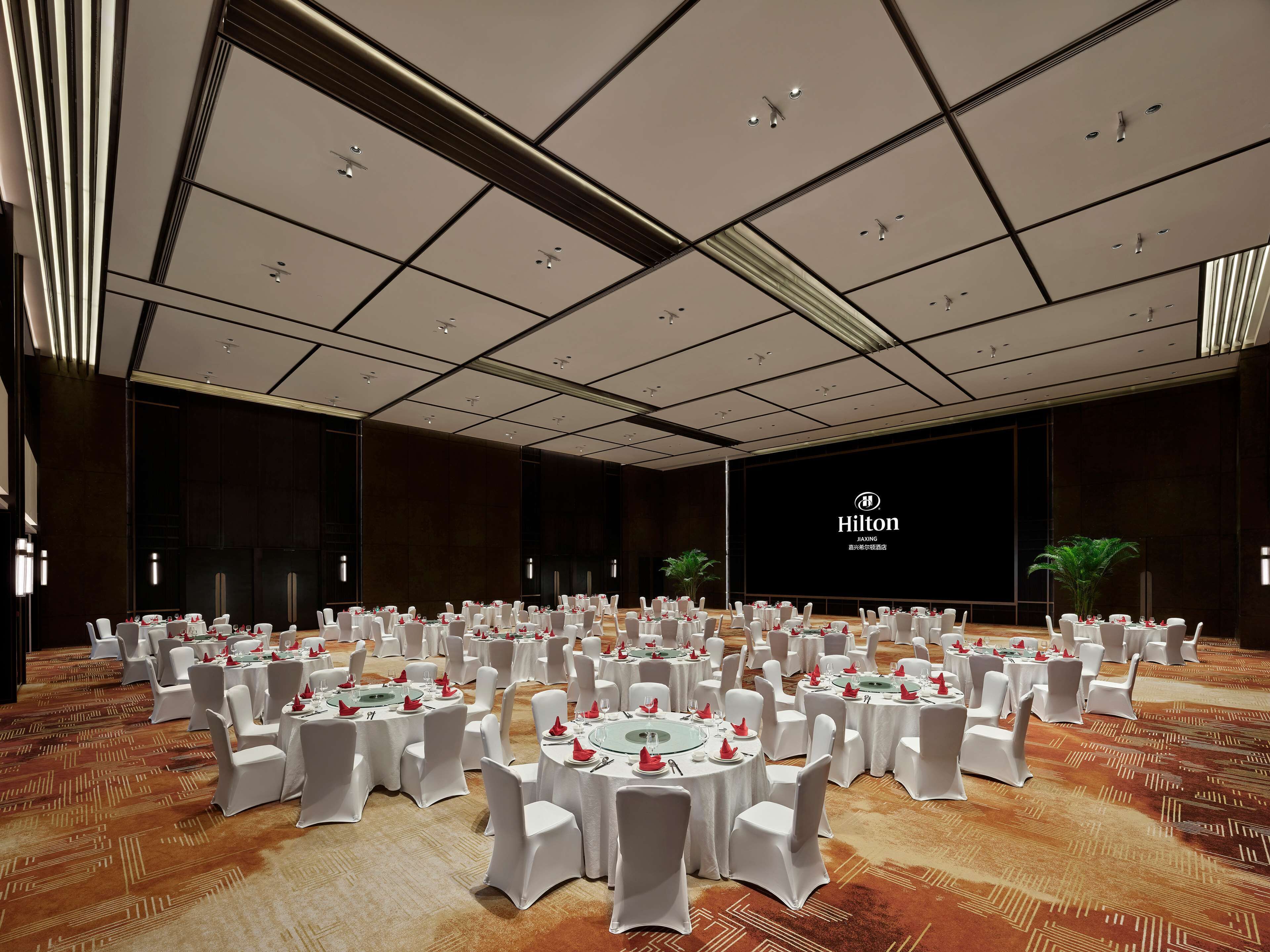 A large, well-lit conference room with multiple round tables set for a formal event, featuring a prominent Hilton logo on the wall.