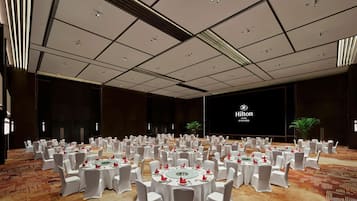 A large, well-lit conference room with multiple round tables set for a formal event, featuring a prominent Hilton logo on the wall.
