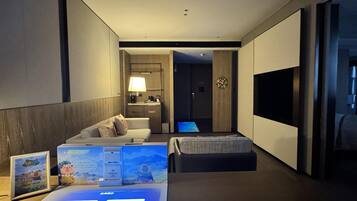 Ein modernes Hotelzimmer mit einer Couch, einem kleinen Bett, einem Schreibtisch mit einem Computer, einem Fernseher und einem sichtbaren Badezimmer im Hintergrund.