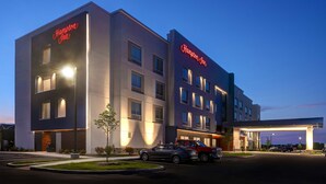 A modern hotel with a prominent "Hampton Inn" sign, a well-lit entrance, and a parking area with several vehicles.