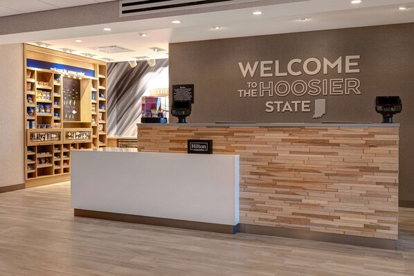 A hotel reception area with a 'Welcome to the Hoosier State' sign, a reception desk, and a shoe display.