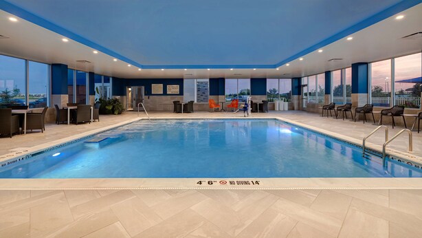 An indoor swimming pool with clear water, surrounded by lounge chairs and tables, and large windows offering views of the cityscape.