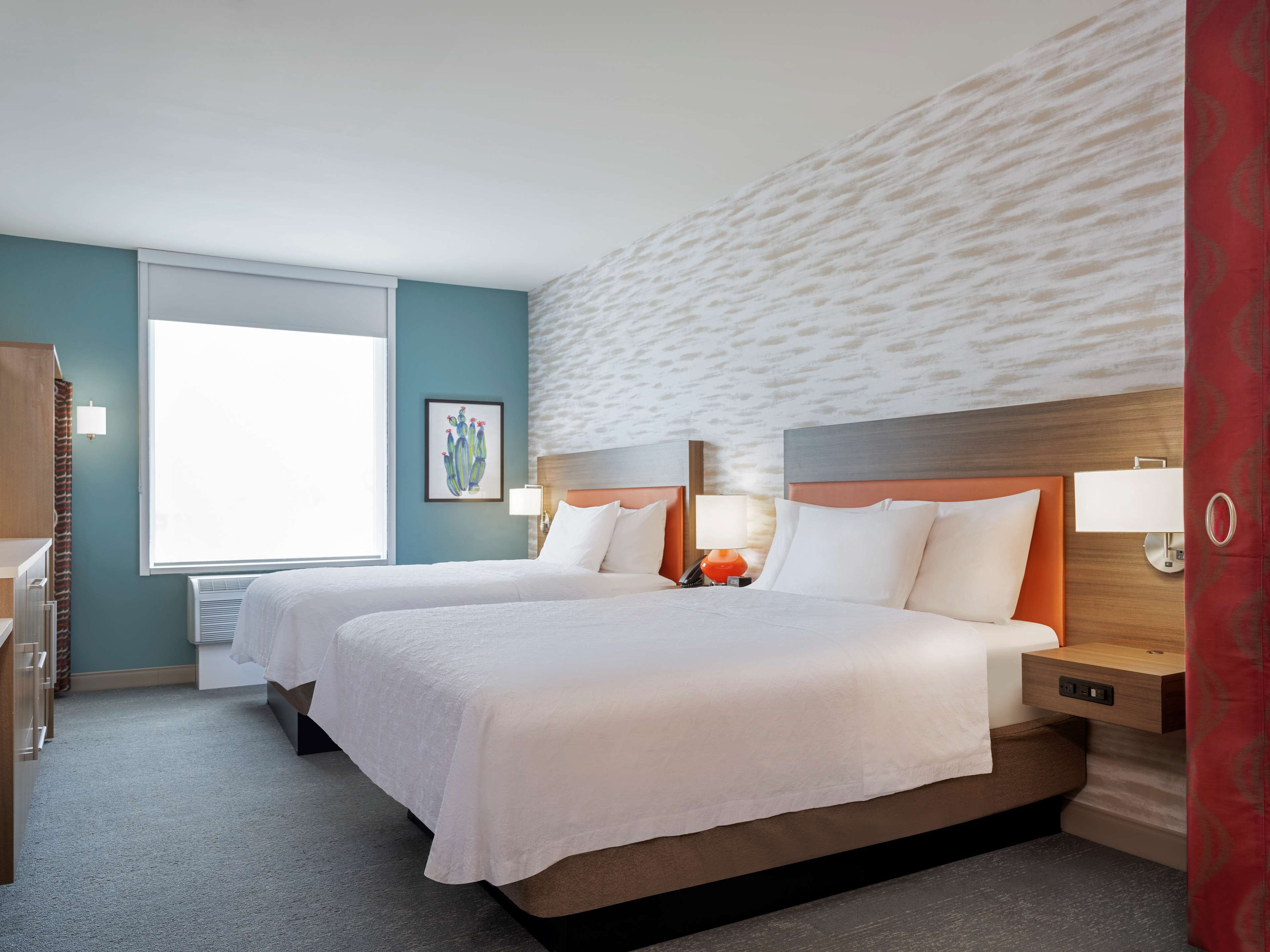 A hotel room with two beds, a window, a wall-mounted artwork, and a red curtain.