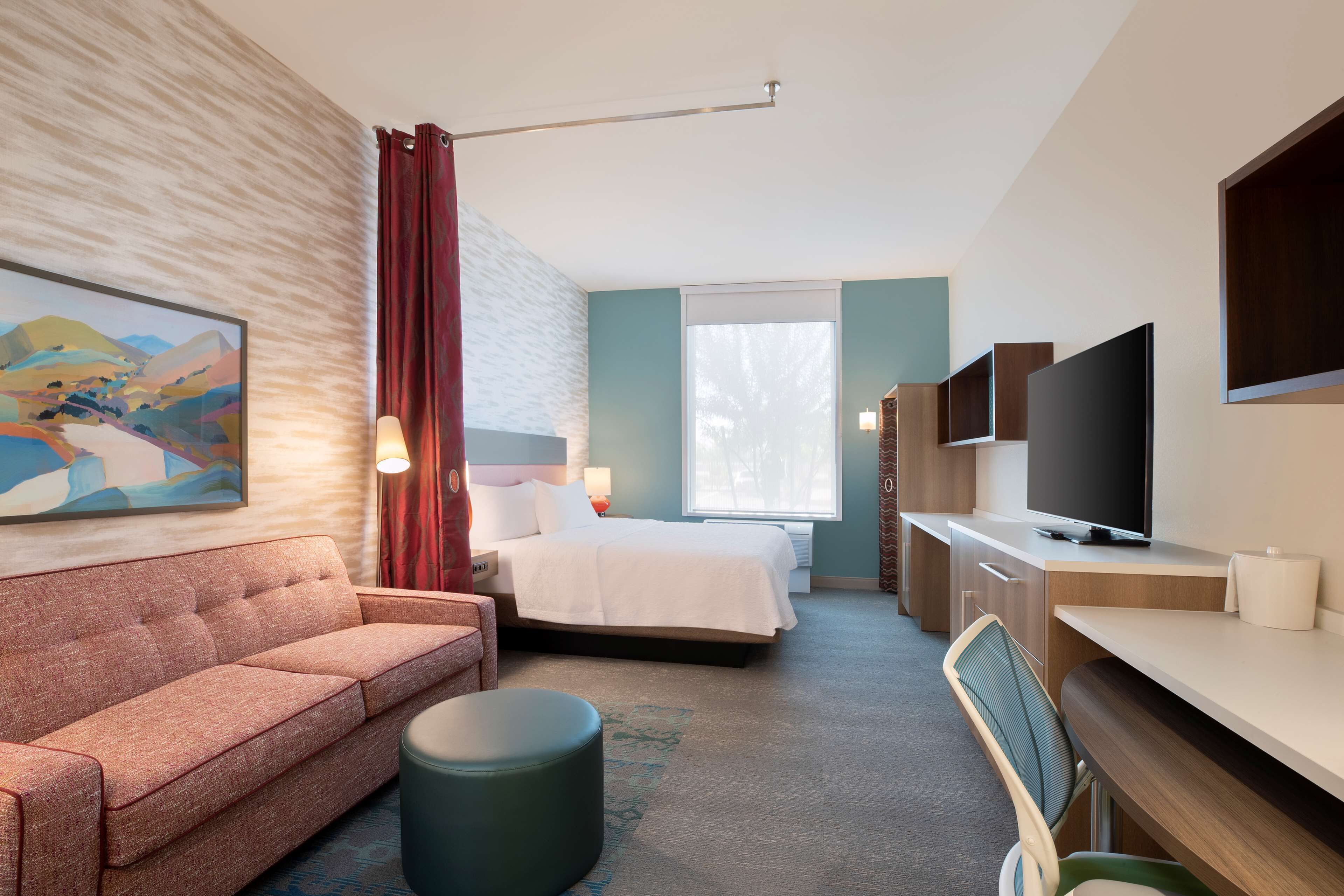 A modern hotel room with a bed, a sofa, a desk, a television, and a painting on the wall.