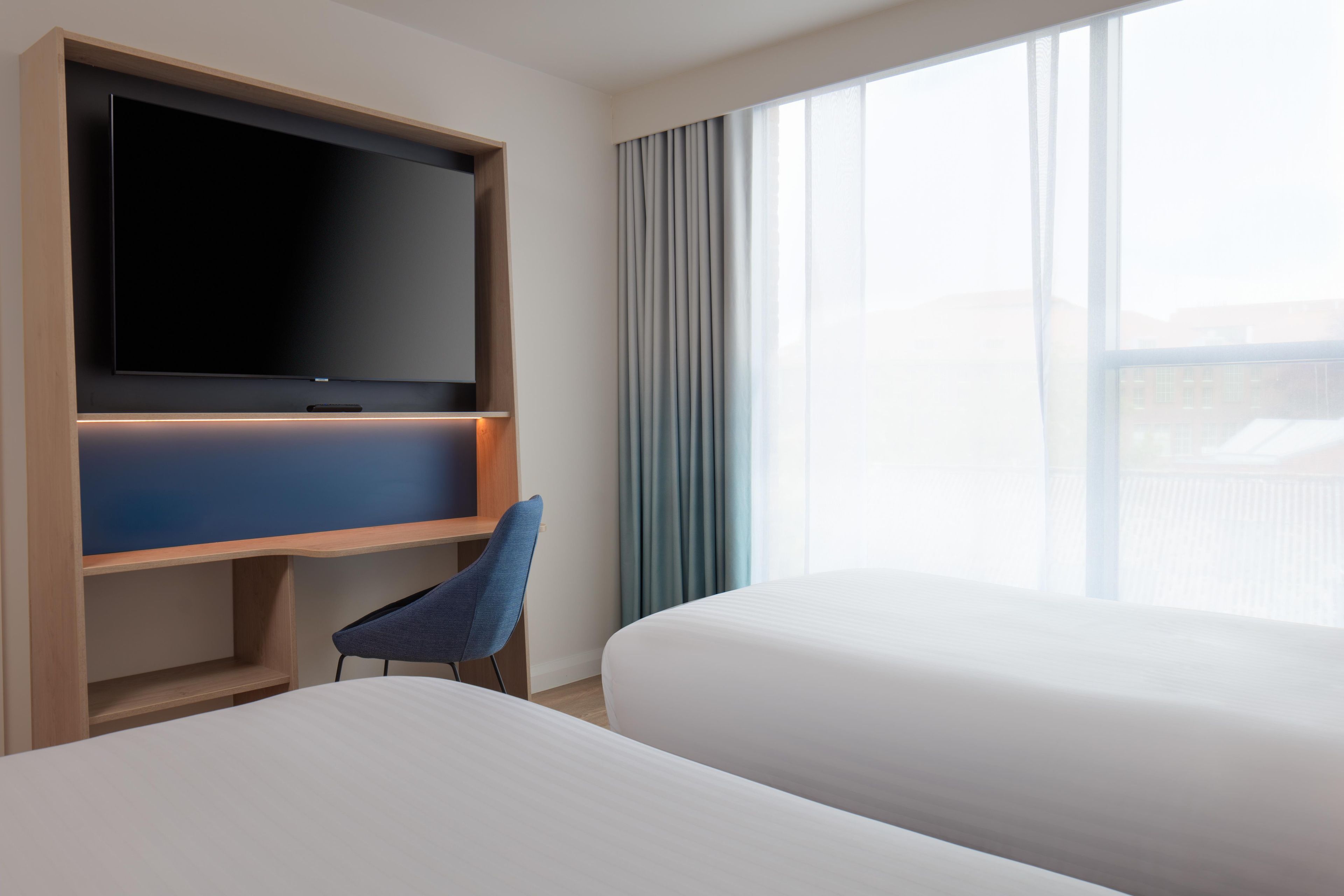 Une chambre d’hôtel équipée d’un téléviseur à écran plat fixé sur un meuble en bois, d’une chaise bleue et de deux lits.