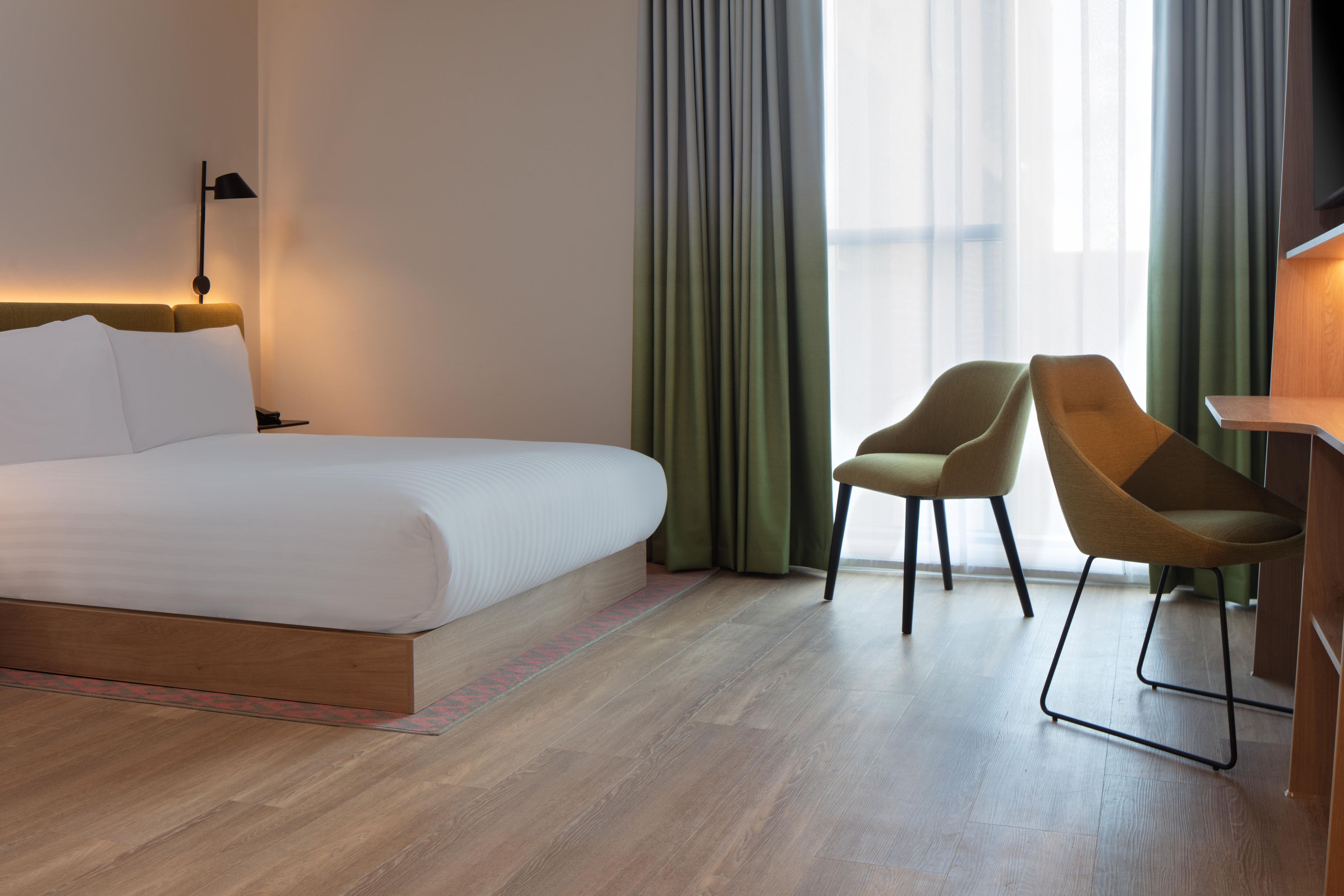 Quarto de hotel com uma cama grande, duas cadeiras, uma escrivaninha e vista para a cidade através de cortinas translúcidas.
