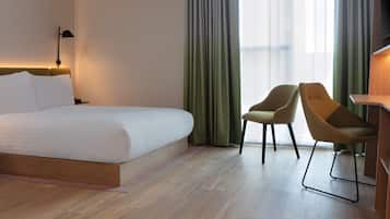Quarto de hotel com uma cama grande, duas cadeiras, uma escrivaninha e vista para a cidade através de cortinas translúcidas.