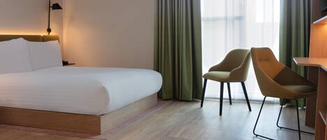 A hotel room with a large bed, two chairs, a desk, and a view of the city through sheer curtains.