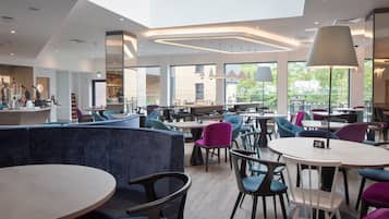 A modern cafe with round tables and chairs, a high ceiling, and large windows offering views of the outdoors.