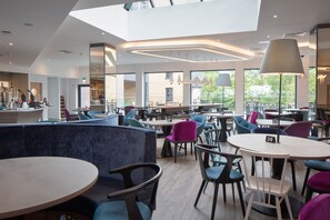 A modern cafe with round tables and chairs, a high ceiling, and large windows offering views of the outdoors. - Hampton by Hilton York Piccadilly (York)