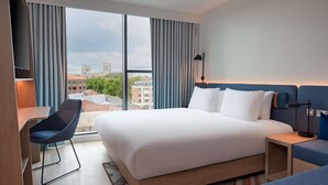 A hotel room with a large bed, a desk, and a chair. There is a view of buildings through the window.